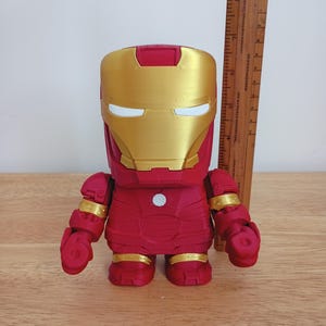 Peut inclure: Figurine d'Iron Man rouge et or. La figurine a un casque et des accents dor&eacute;s, avec un corps et des membres rouges. La figurine mesure environ 15 cm de haut et se tient sur une surface en bois.