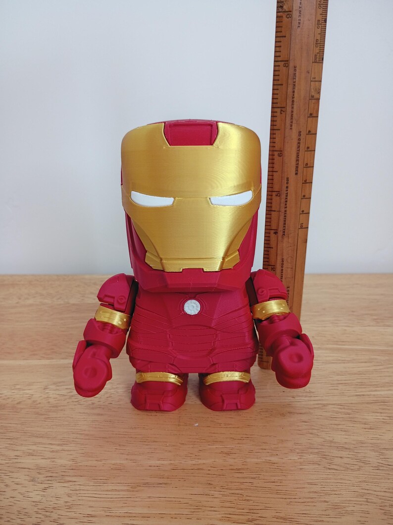 Peut inclure: Figurine d'Iron Man rouge et or. La figurine a un casque et des accents dor&eacute;s, avec un corps et des membres rouges. La figurine mesure environ 15 cm de haut et se tient sur une surface en bois.