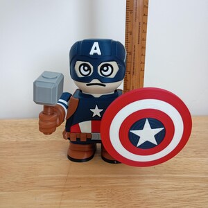 Peut inclure: Figurine de Captain America avec un casque bleu orn&eacute; d'un "A" blanc, une expression s&eacute;rieuse et une &eacute;toile sur la poitrine. La figurine tient un marteau gris et un bouclier rouge, blanc et bleu. Une r&egrave;gle en bois est en arri&egrave;re-plan.
