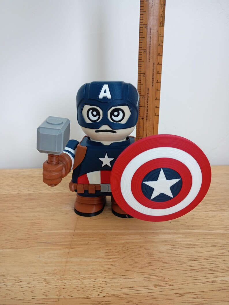 Peut inclure: Figurine de Captain America avec un casque bleu orn&eacute; d'un "A" blanc, une expression s&eacute;rieuse et une &eacute;toile sur la poitrine. La figurine tient un marteau gris et un bouclier rouge, blanc et bleu. Une r&egrave;gle en bois est en arri&egrave;re-plan.