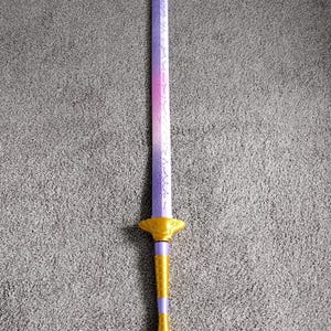May include: A decorative sword with a purple and pink blade, transitioning to a gold and purple hilt. The blade has etched designs. The hilt features a gold guard and a detailed handle. The sword is set against a gray background.