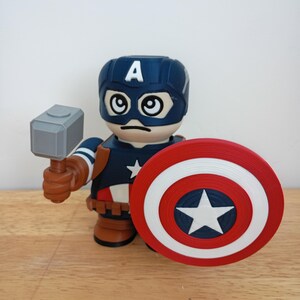 Peut inclure: Figurine de Captain America avec un casque bleu orn&eacute; d'un "A" blanc, une expression s&eacute;v&egrave;re et de grands yeux. La figurine tient un marteau gris et un bouclier rouge, blanc et bleu avec une &eacute;toile blanche. La figurine est sur une surface en bois.