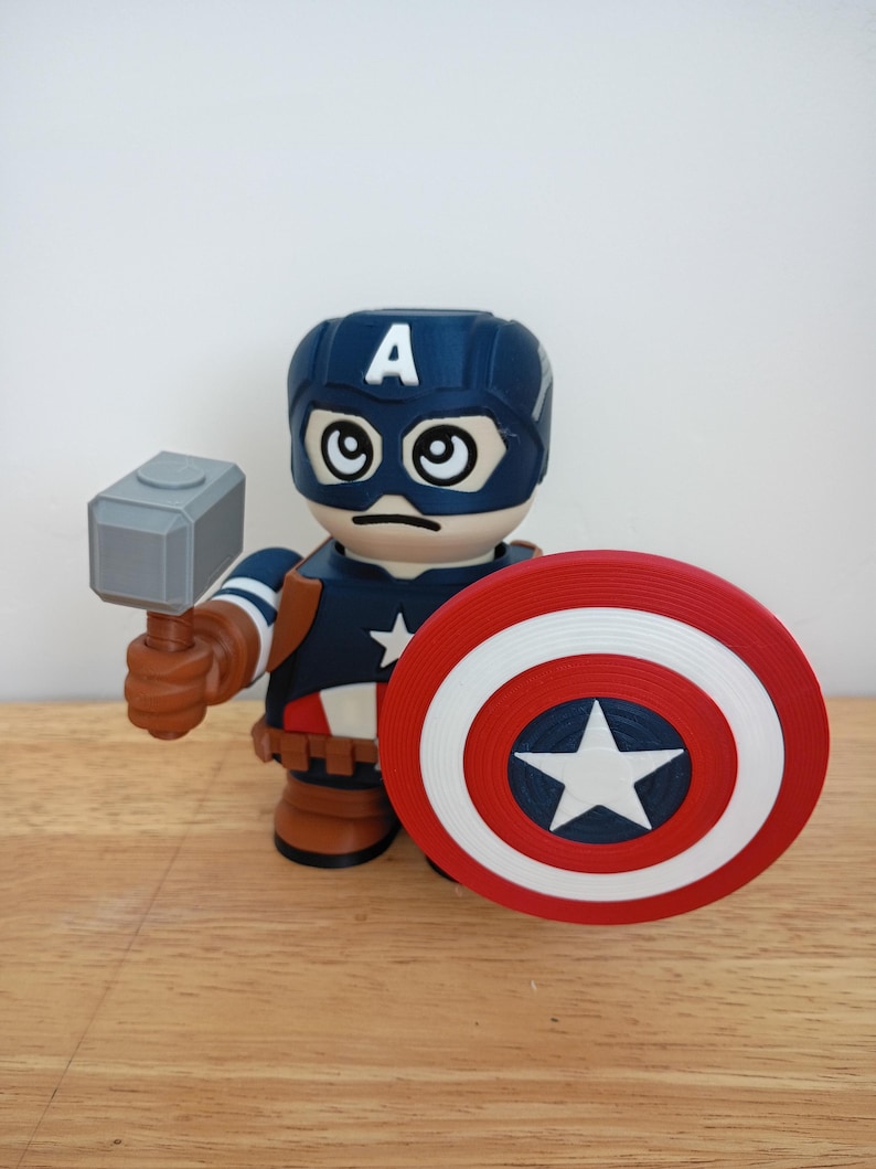 Peut inclure: Figurine de Captain America avec un casque bleu orn&eacute; d'un "A" blanc, une expression s&eacute;v&egrave;re et de grands yeux. La figurine tient un marteau gris et un bouclier rouge, blanc et bleu avec une &eacute;toile blanche. La figurine est sur une surface en bois.