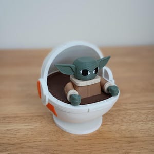May include: A figurine of a green-skinned character with large ears, wearing a brown coat, seated in a white pod with orange accents. The pod has a brown interior. The figurine is a popular character from a science fiction franchise.