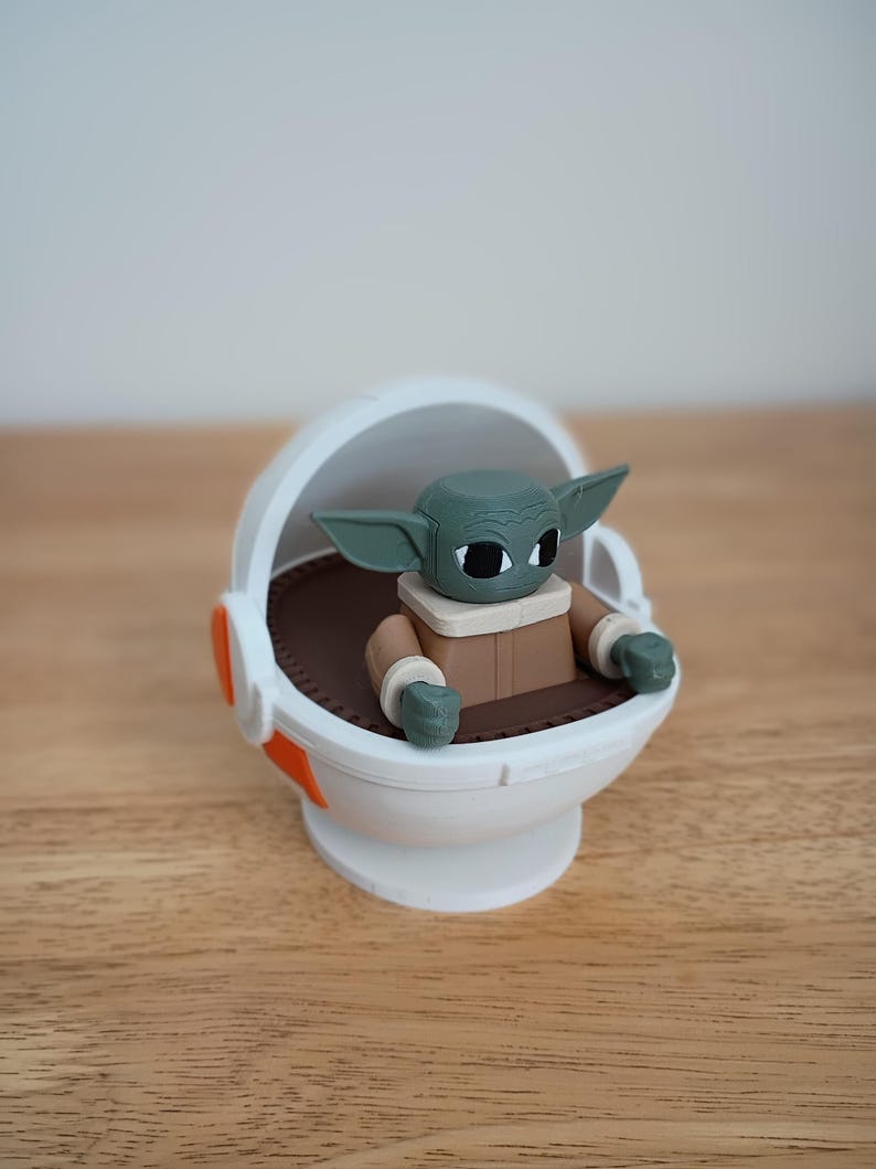 May include: A figurine of a green-skinned character with large ears, wearing a brown coat, seated in a white pod with orange accents. The pod has a brown interior. The figurine is a popular character from a science fiction franchise.
