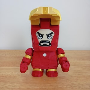 Peut inclure: Figurine rouge d'Iron Man avec des d&eacute;tails dor&eacute;s sur les articulations et le casque. La figurine a une t&ecirc;te carr&eacute;e avec une expression s&eacute;v&egrave;re et une pi&egrave;ce de casque dor&eacute;e. La figurine est pos&eacute;e sur une surface en bois.
