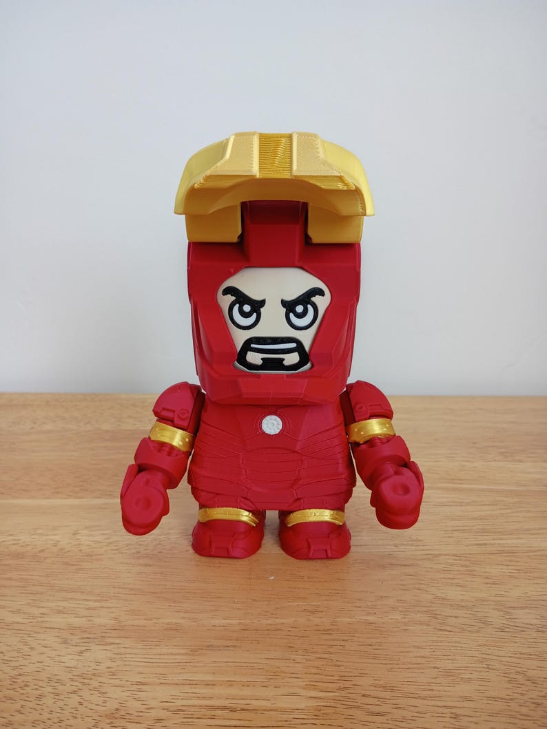 Peut inclure: Figurine rouge d'Iron Man avec des d&eacute;tails dor&eacute;s sur les articulations et le casque. La figurine a une t&ecirc;te carr&eacute;e avec une expression s&eacute;v&egrave;re et une pi&egrave;ce de casque dor&eacute;e. La figurine est pos&eacute;e sur une surface en bois.
