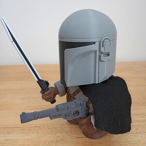 May include: A gray Mandalorian action figure with a black cape, holding a silver and black lightsaber and a gray blaster pistol. The figure has a gray helmet and brown boots. The figure is on a wooden surface.