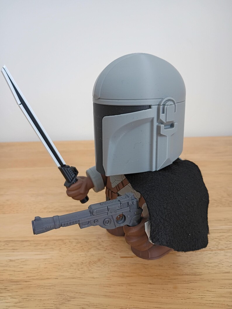 May include: A gray Mandalorian action figure with a black cape, holding a silver and black lightsaber and a gray blaster pistol. The figure has a gray helmet and brown boots. The figure is on a wooden surface.