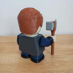 Peut inclure: Une figurine imprim&eacute;e en 3D repr&eacute;sentant une personne aux cheveux bruns, v&ecirc;tue d'une veste gris fonc&eacute; et d'un pantalon bleu. La figurine tient une petite hache grise et marron. La figurine est pos&eacute;e sur une surface en bois.