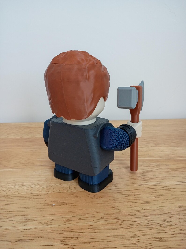 Peut inclure: Une figurine imprim&eacute;e en 3D repr&eacute;sentant une personne aux cheveux bruns, v&ecirc;tue d'une veste gris fonc&eacute; et d'un pantalon bleu. La figurine tient une petite hache grise et marron. La figurine est pos&eacute;e sur une surface en bois.