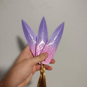 Könnte beinhalten: Drei lila dekorative Klingen mit aufwendigen Designs und einem Farbverlauf, die von einer goldenen Quaste zusammengehalten werden. Die Klingen haben eine spitze Form und ein detailliertes Muster auf ihrer Oberfläche.