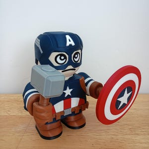 Peut inclure: Figurine imprim&eacute;e en 3D de Captain America tenant un marteau gris et un bouclier rouge, blanc et bleu. La figurine porte un casque bleu avec un "A" blanc et un uniforme bleu marine et blanc avec une &eacute;toile blanche.