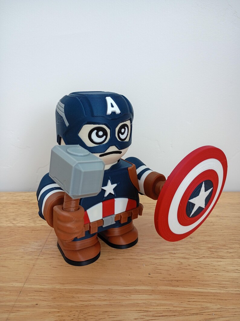 Peut inclure: Figurine imprim&eacute;e en 3D de Captain America tenant un marteau gris et un bouclier rouge, blanc et bleu. La figurine porte un casque bleu avec un "A" blanc et un uniforme bleu marine et blanc avec une &eacute;toile blanche.