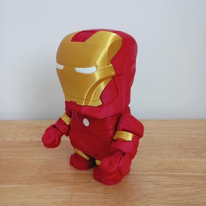 Peut inclure: Figurine d'Iron Man imprim&eacute;e en 3D, de style chibi. La figurine est principalement rouge avec des accents dor&eacute;s sur le casque et les articulations. Elle a un corps rond, de petits bras et de grands pieds, ce qui lui donne un aspect de dessin anim&eacute;.