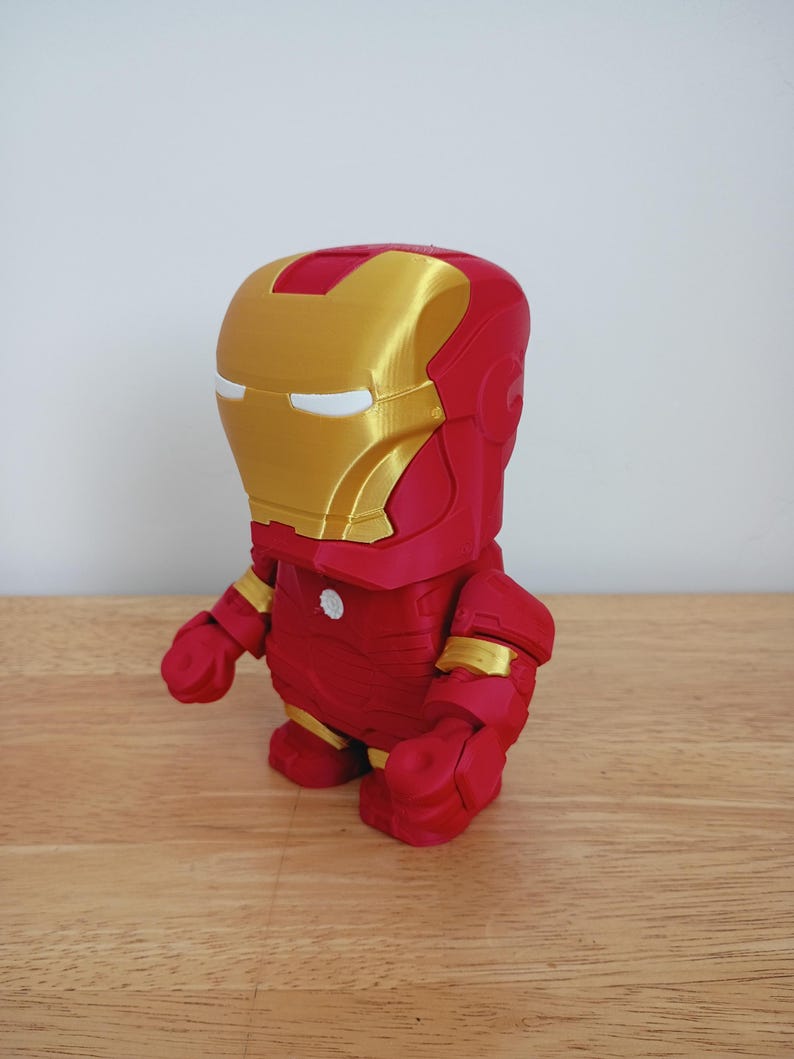 Peut inclure: Figurine d'Iron Man imprim&eacute;e en 3D, de style chibi. La figurine est principalement rouge avec des accents dor&eacute;s sur le casque et les articulations. Elle a un corps rond, de petits bras et de grands pieds, ce qui lui donne un aspect de dessin anim&eacute;.