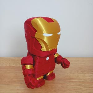 Peut inclure: Figurine d'Iron Man rouge et or. La figurine a un corps rouge avec des accents dor&eacute;s sur le casque, les &eacute;paules et les mains. Les yeux sont blancs. La figurine est pos&eacute;e sur une surface en bois.