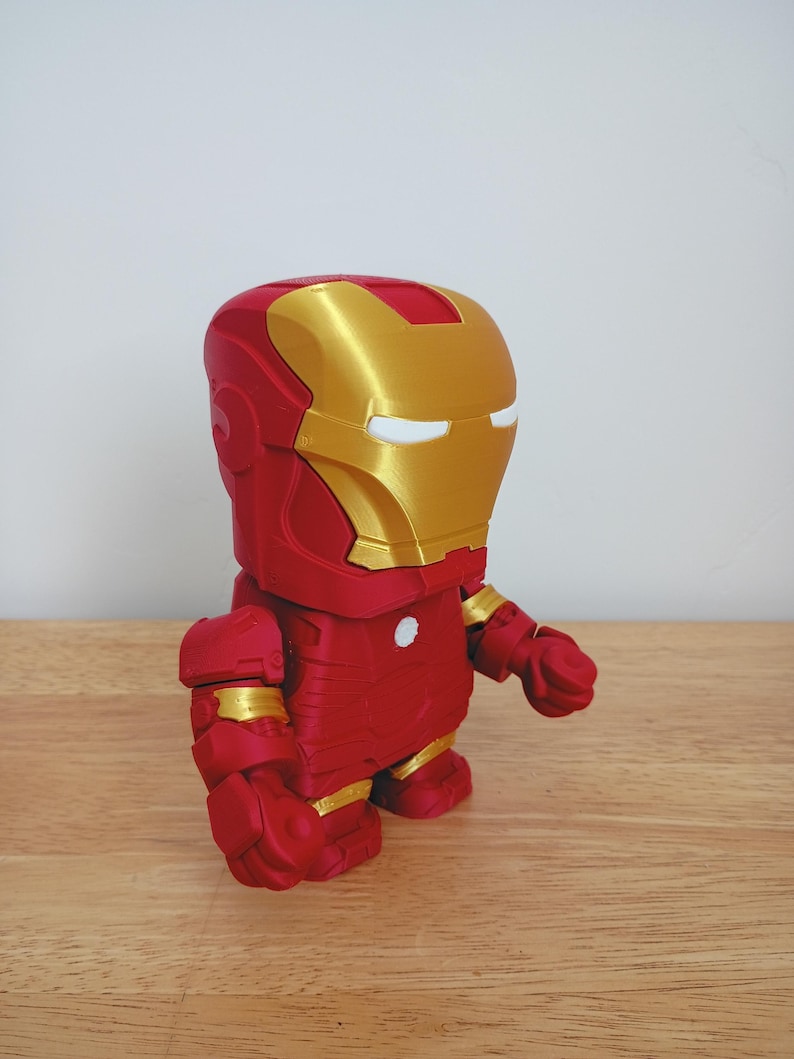 Peut inclure: Figurine d'Iron Man rouge et or. La figurine a un corps rouge avec des accents dor&eacute;s sur le casque, les &eacute;paules et les mains. Les yeux sont blancs. La figurine est pos&eacute;e sur une surface en bois.