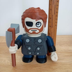 Peut inclure: Une figurine de Thor avec une barbe brune, un cache-&oelig;il et des cheveux roux, tenant une hache grise et marron. La figurine porte une tenue grise et bleue. Une r&egrave;gle en bois est &agrave; c&ocirc;t&eacute; de la figurine.