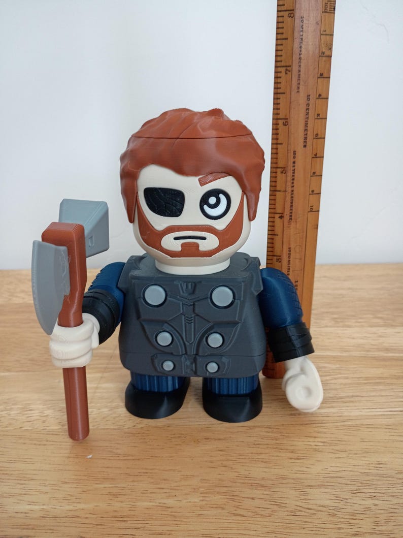 Peut inclure: Une figurine de Thor avec une barbe brune, un cache-&oelig;il et des cheveux roux, tenant une hache grise et marron. La figurine porte une tenue grise et bleue. Une r&egrave;gle en bois est &agrave; c&ocirc;t&eacute; de la figurine.