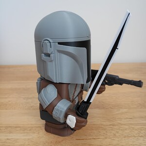 May include: A gray and brown Mandalorian figure holding a black and white sword and a black blaster pistol. The figure has a gray helmet and armor, with brown and gray details. The figure is standing on a wooden surface.