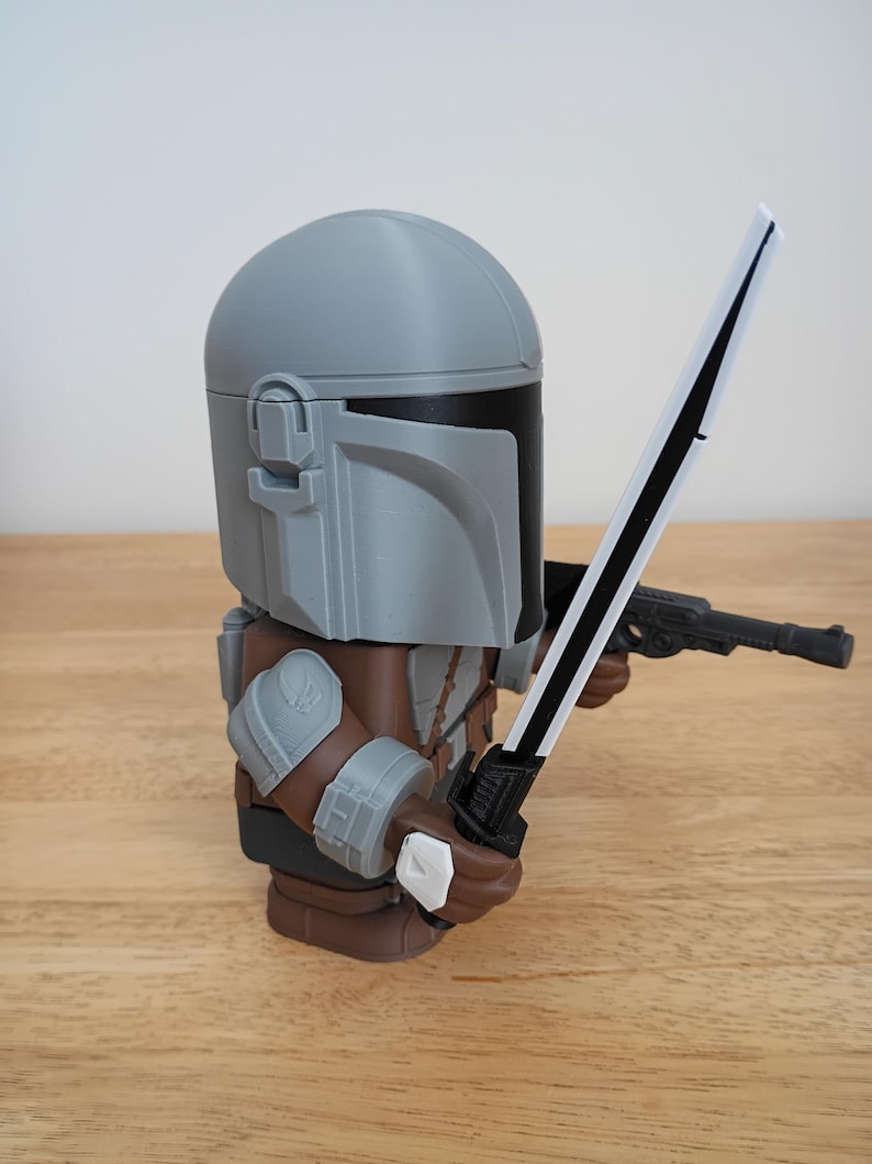 May include: A gray and brown Mandalorian figure holding a black and white sword and a black blaster pistol. The figure has a gray helmet and armor, with brown and gray details. The figure is standing on a wooden surface.