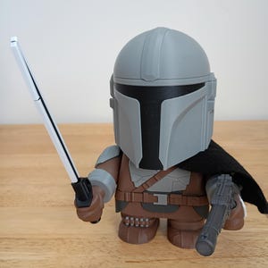 May include: A stylized figurine of the Mandalorian character from Star Wars. The figure is wearing a gray helmet and armor, a black cape, and holding a white and black sword and a blaster. The figure is brown, gray, and black.