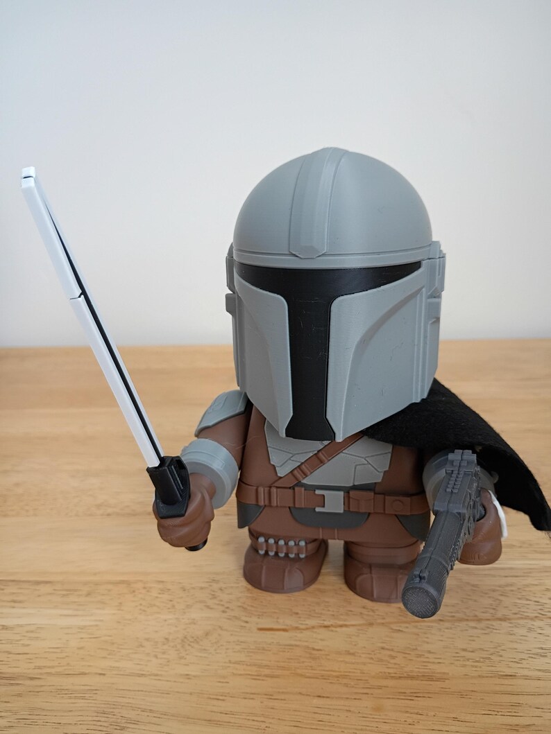 May include: A stylized figurine of the Mandalorian character from Star Wars. The figure is wearing a gray helmet and armor, a black cape, and holding a white and black sword and a blaster. The figure is brown, gray, and black.