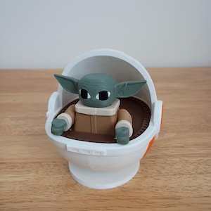 May include: A green-skinned Grogu figure in a white and orange carrier. The figure has large black eyes and pointed ears. The carrier is white with brown and orange accents, sitting on a wooden surface.