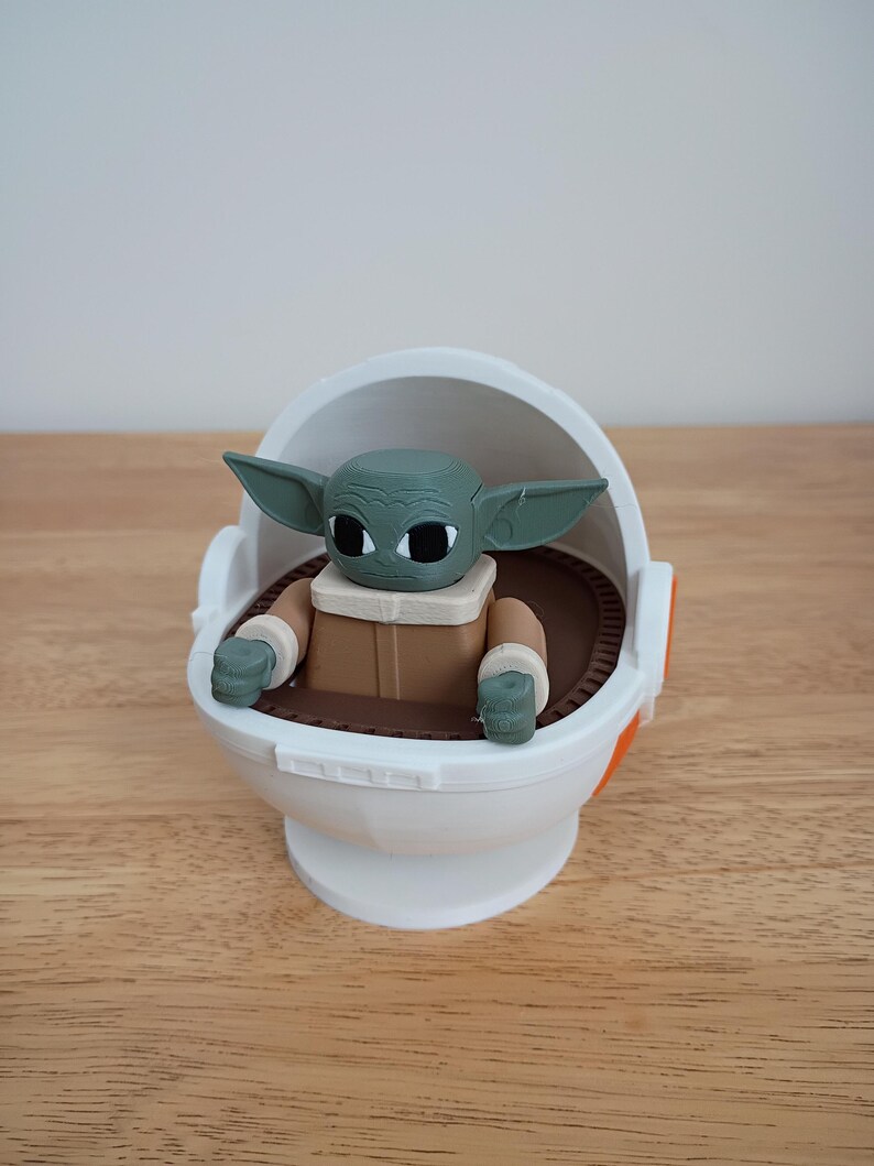 May include: A green-skinned Grogu figure in a white and orange carrier. The figure has large black eyes and pointed ears. The carrier is white with brown and orange accents, sitting on a wooden surface.
