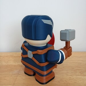 Peut inclure: Figurine imprim&eacute;e en 3D de Captain America, avec un casque bleu et une aile blanche, des sangles marron et un bouclier rouge. La figurine tient un marteau gris avec un manche marron. Pos&eacute;e sur une surface en bois.