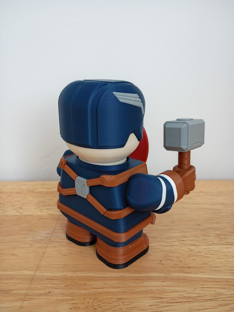 Peut inclure: Figurine imprim&eacute;e en 3D de Captain America, avec un casque bleu et une aile blanche, des sangles marron et un bouclier rouge. La figurine tient un marteau gris avec un manche marron. Pos&eacute;e sur une surface en bois.