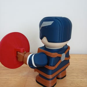 Peut inclure: Figurine imprim&eacute;e en 3D d'un super-h&eacute;ros en uniforme bleu et marron, tenant un bouclier rouge. La figurine a un design stylis&eacute; avec un casque orn&eacute; d'un embl&egrave;me d'aile blanc. La figurine est sur une surface en bois.