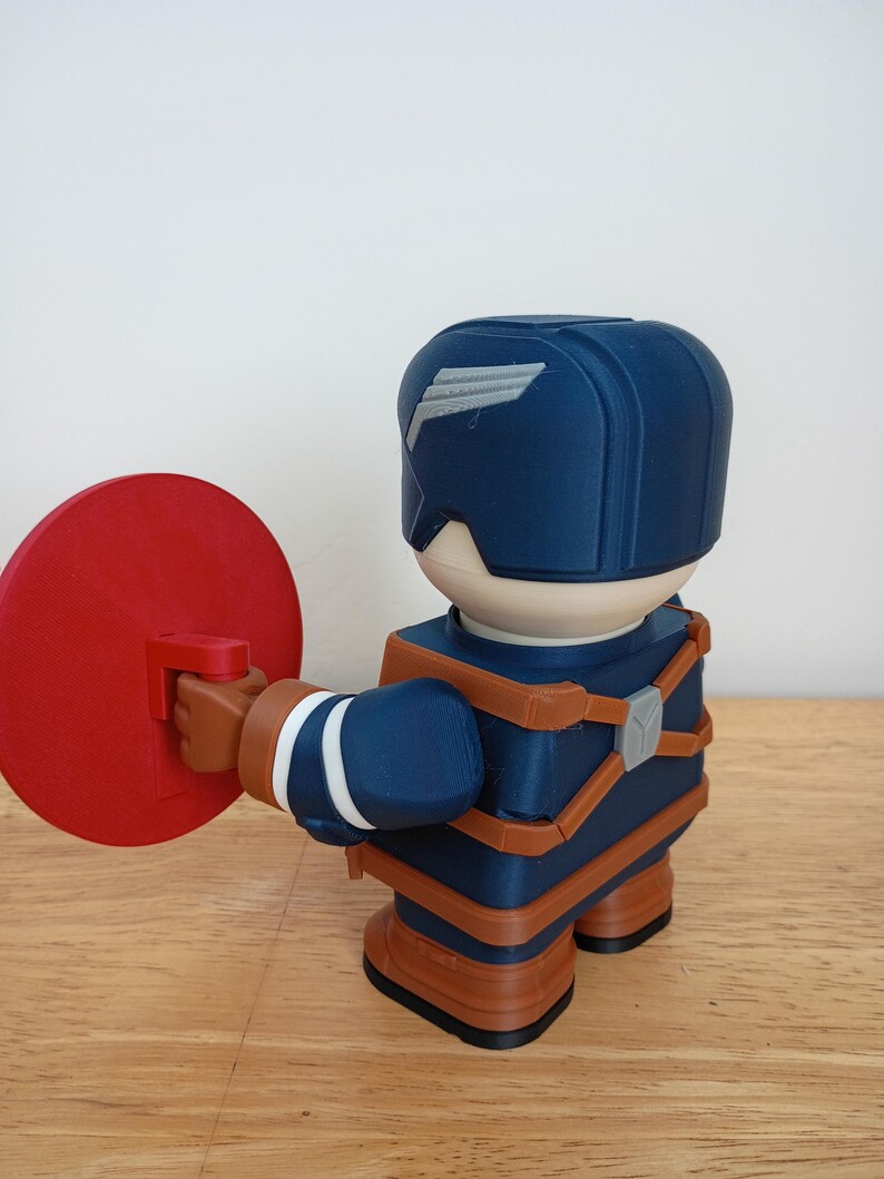 Peut inclure: Figurine imprim&eacute;e en 3D d'un super-h&eacute;ros en uniforme bleu et marron, tenant un bouclier rouge. La figurine a un design stylis&eacute; avec un casque orn&eacute; d'un embl&egrave;me d'aile blanc. La figurine est sur une surface en bois.