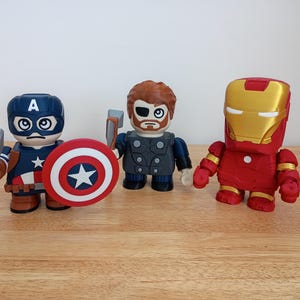 Peut inclure: Trois figurines de super-h&eacute;ros : Captain America avec un bouclier et un marteau, Thor avec un marteau et Iron Man dans une armure rouge et or. Les figurines sont stylis&eacute;es avec de grosses t&ecirc;tes et des corps carr&eacute;s, sur une surface en bois.