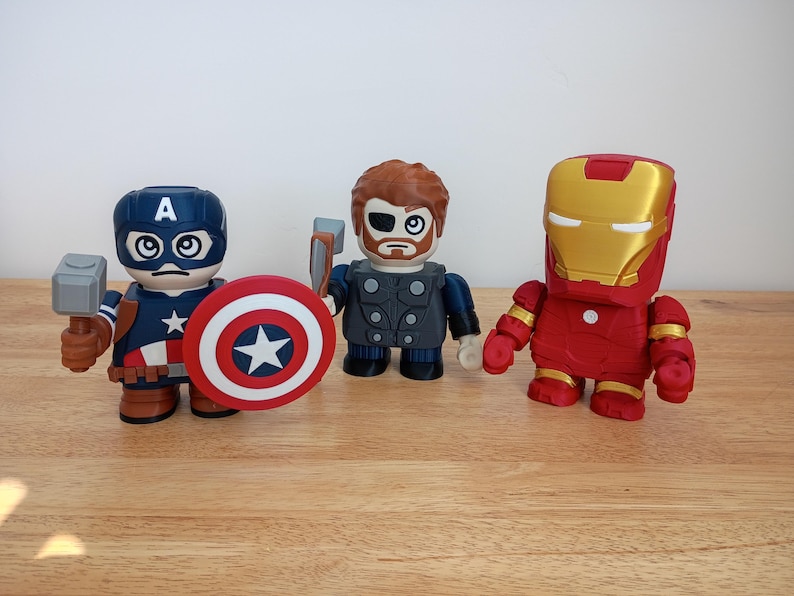 Peut inclure: Trois figurines de super-h&eacute;ros : Captain America avec un bouclier et un marteau, Thor avec un marteau et Iron Man dans une armure rouge et or. Les figurines sont stylis&eacute;es avec de grosses t&ecirc;tes et des corps carr&eacute;s, sur une surface en bois.