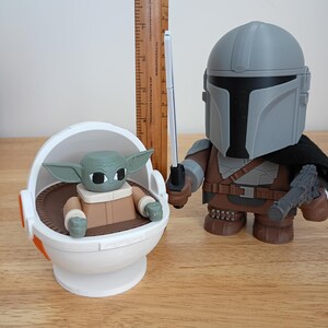 May include: A pair of Star Wars themed figurines. One is a Mandalorian figure with a blaster and a lightsaber. The other is a Grogu figure in a floating pod. The figures are on a wooden surface.