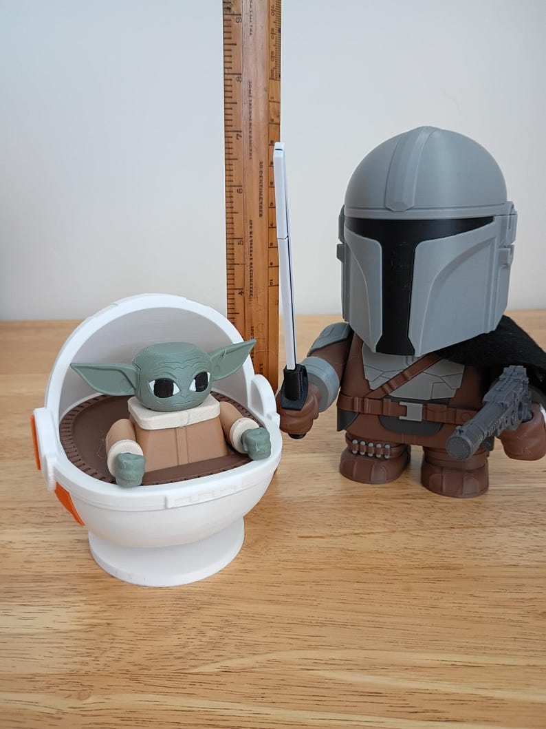 May include: A pair of Star Wars themed figurines. One is a Mandalorian figure with a blaster and a lightsaber. The other is a Grogu figure in a floating pod. The figures are on a wooden surface.