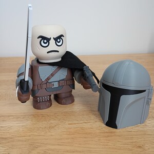May include: A stylized figure of a character from a science fiction series, with a stern expression, holding a silver sword and a blaster. A gray helmet is placed next to the figure. The figure is wearing brown and gray armor with a black cape.