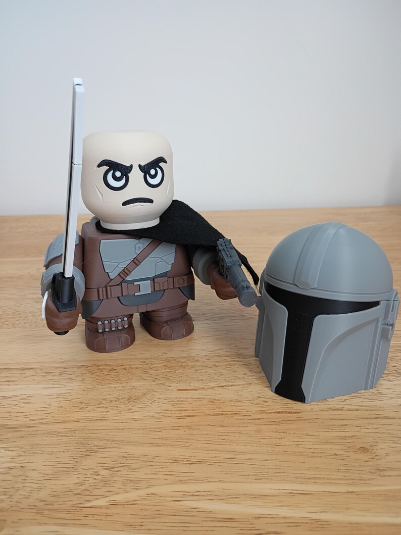 May include: A stylized figure of a character from a science fiction series, with a stern expression, holding a silver sword and a blaster. A gray helmet is placed next to the figure. The figure is wearing brown and gray armor with a black cape.