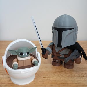 May include: A pair of Star Wars-themed figurines. One is a Mandalorian figure in gray and brown armor, holding a blaster and a lightsaber. The other is a Grogu figure in a white and brown carrier.