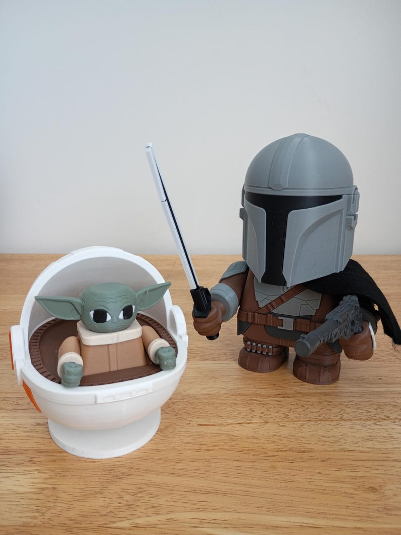 May include: A pair of Star Wars-themed figurines. One is a Mandalorian figure in gray and brown armor, holding a blaster and a lightsaber. The other is a Grogu figure in a white and brown carrier.