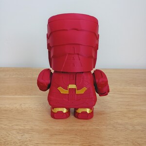 Peut inclure: Figurine rouge d'un super-h&eacute;ros, vue de dos. La figurine pr&eacute;sente un design anguleux avec des d&eacute;tails dor&eacute;s sur la poitrine et les pieds. Elle est pos&eacute;e sur une surface en bois.