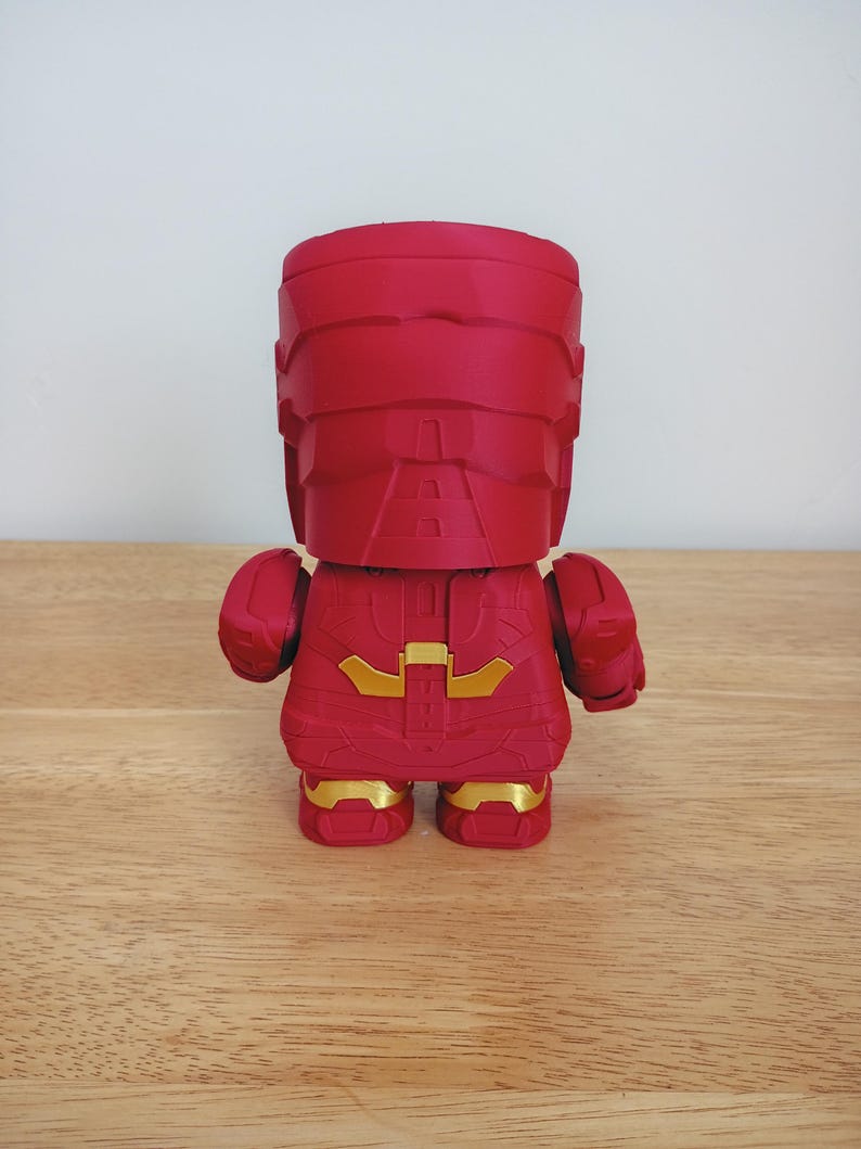 Peut inclure: Figurine rouge d'un super-h&eacute;ros, vue de dos. La figurine pr&eacute;sente un design anguleux avec des d&eacute;tails dor&eacute;s sur la poitrine et les pieds. Elle est pos&eacute;e sur une surface en bois.