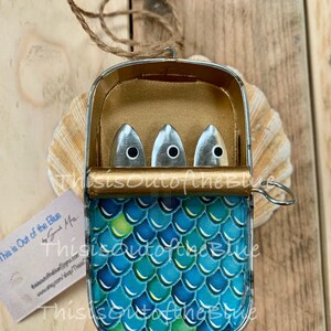 May include: A sardine can-shaped ornament with a blue and green scale design. The can is open, showing three silver fish with black eyes. A small tag with text is visible. It hangs from a twine loop.