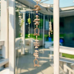 Può includere: Mobile sospeso in legno e perline. Il mobile presenta pezzi di legno naturale, piccole perline blu e due elementi in iuta a forma di campana. Il design è sospeso da un cordoncino naturale, creando un'estetica costiera o bohémien.