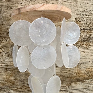 Peut inclure: Un carillon à vent en coquillages blancs avec un sommet en bois. Les coquillages sont disposés en cascade et sont suspendus au sommet par des cordes. Le carillon à vent est suspendu à une surface en bois.