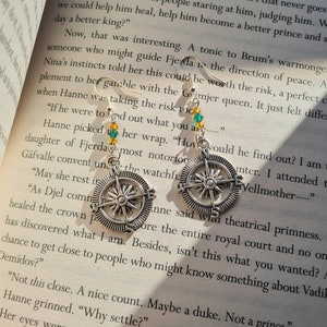 Puede incluir: Un par de pendientes de brújula plateados con cuentas de vidrio verde y azul. Los pendientes cuelgan de un libro con texto sobre una reina ficticia.