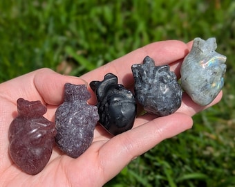 May include: Five carved gemstone heart sculptures in varying colors: red, purple, black, gray, and iridescent. The heart-shaped stones are held in an open hand against a backdrop of green grass.