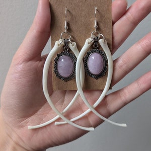Peut inclure: Une paire de boucles d'oreilles pendantes uniques. Chaque boucle d'oreille présente une pierre ovale violet clair sertie dans un cadre décoratif argenté, suspendue à un crochet argenté. La pierre est fixée à une structure incurvée en forme d'os.